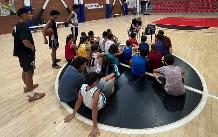 Entrenador explicant un exercici a un grup de jugadors asseguts a la pista, al campus del CB Grup Barna