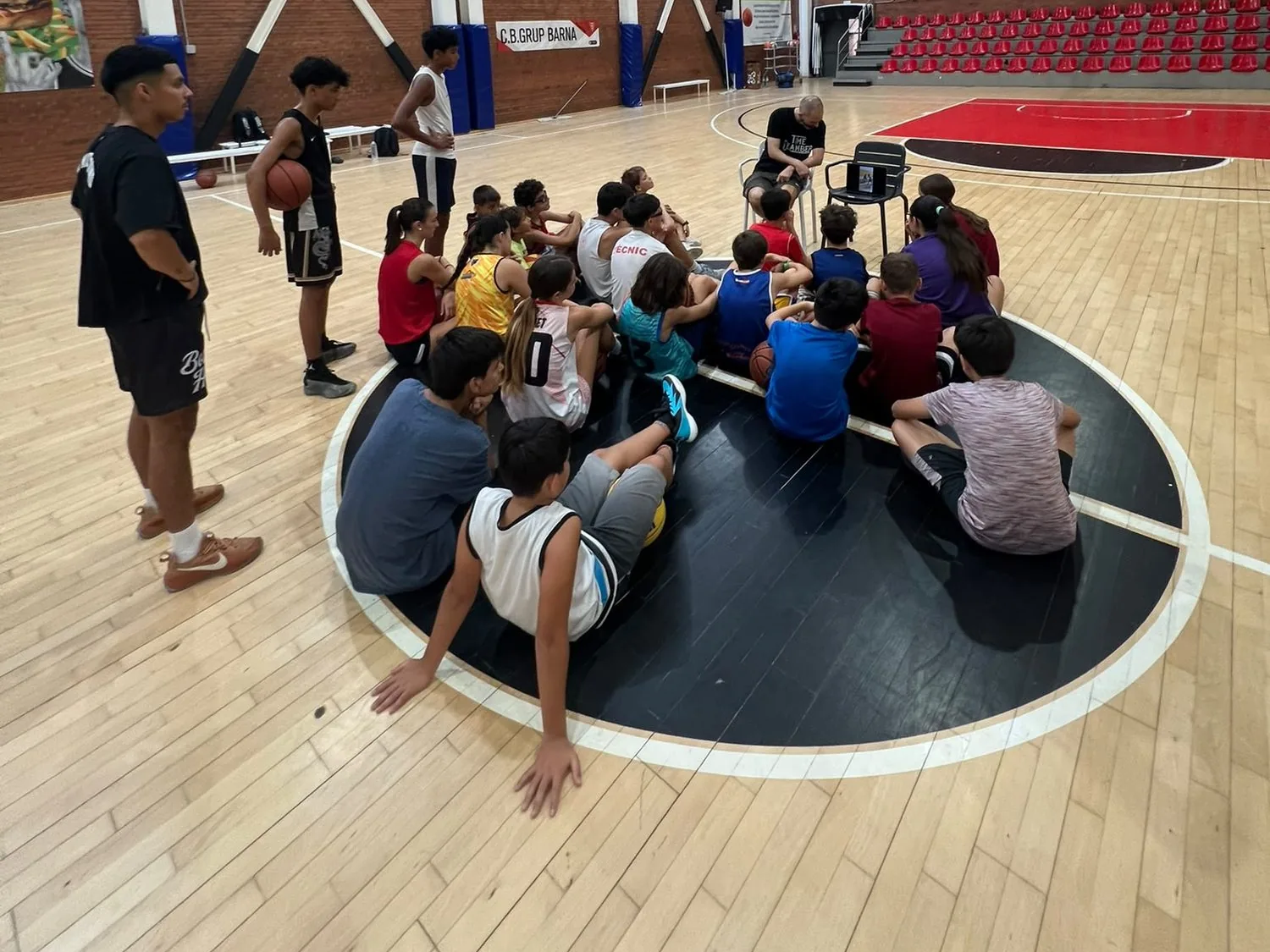Entrenador explicant un exercici a un grup de jugadors asseguts a la pista, al campus del CB Grup Barna